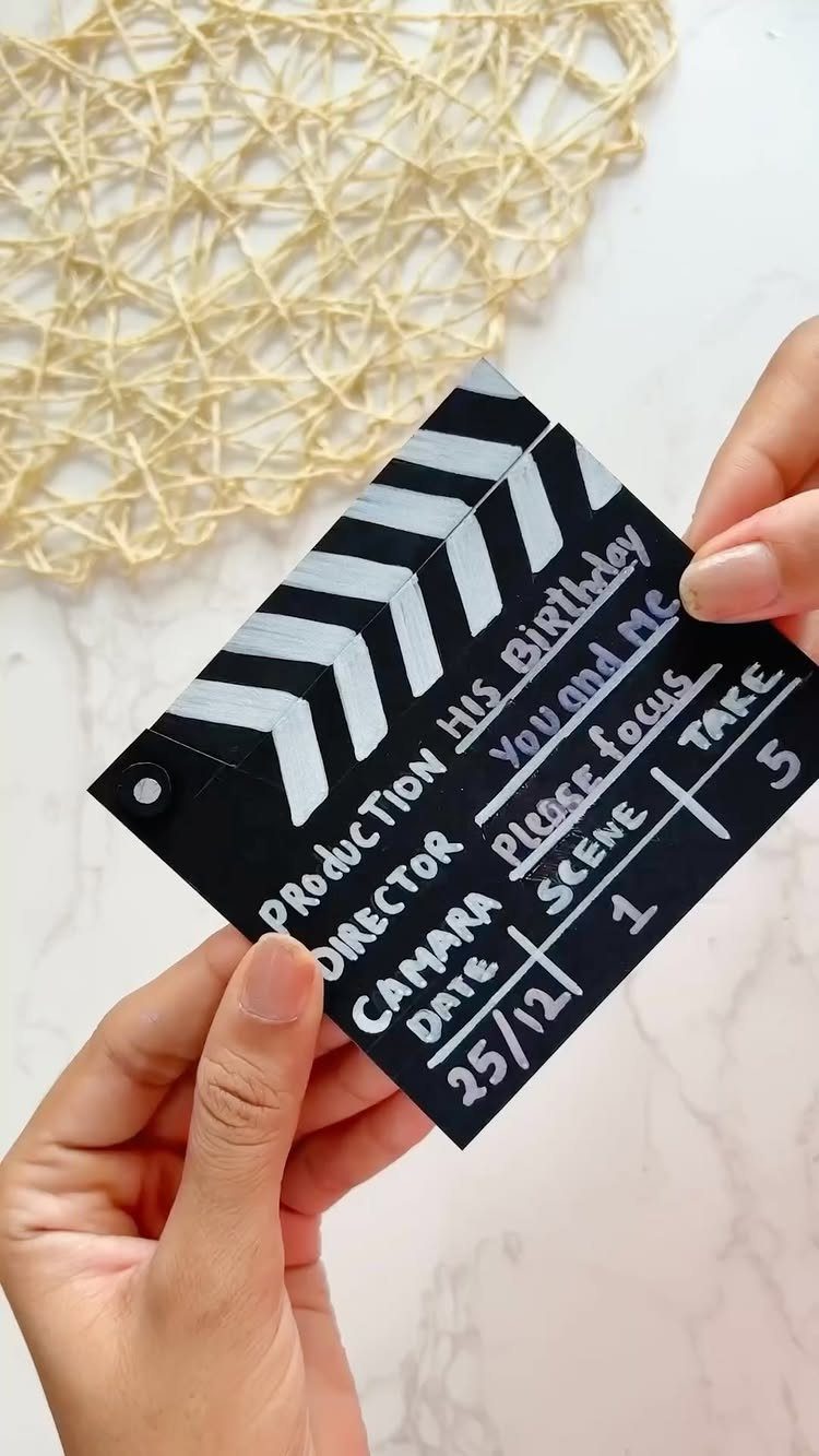 Hands holding a movie clapperboard card with handwritten birthday messages on marble surface