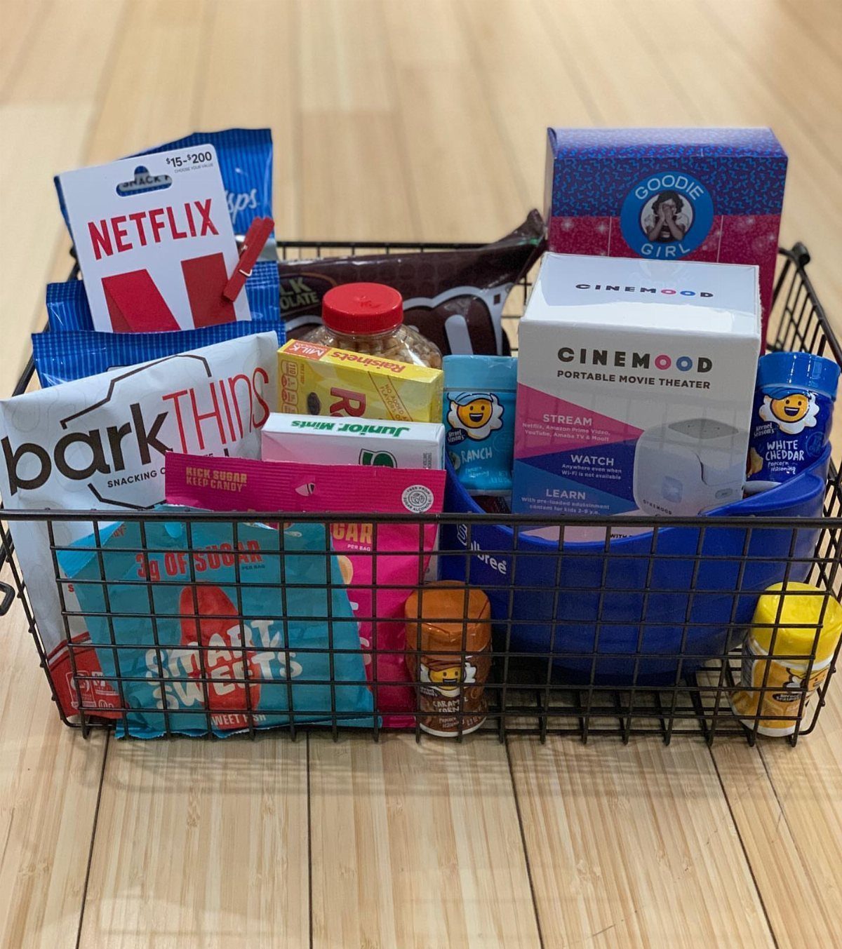 Movie-themed gift basket with Netflix card, CineMood projector, and various snacks on wood floor.