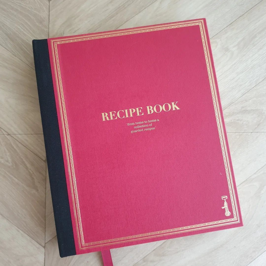 Red hardcover recipe book with gold lettering resting on a light wooden floor.