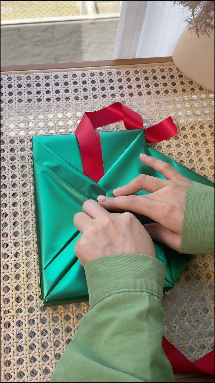 Hands wrapping a gift in shiny green paper with a red ribbon.