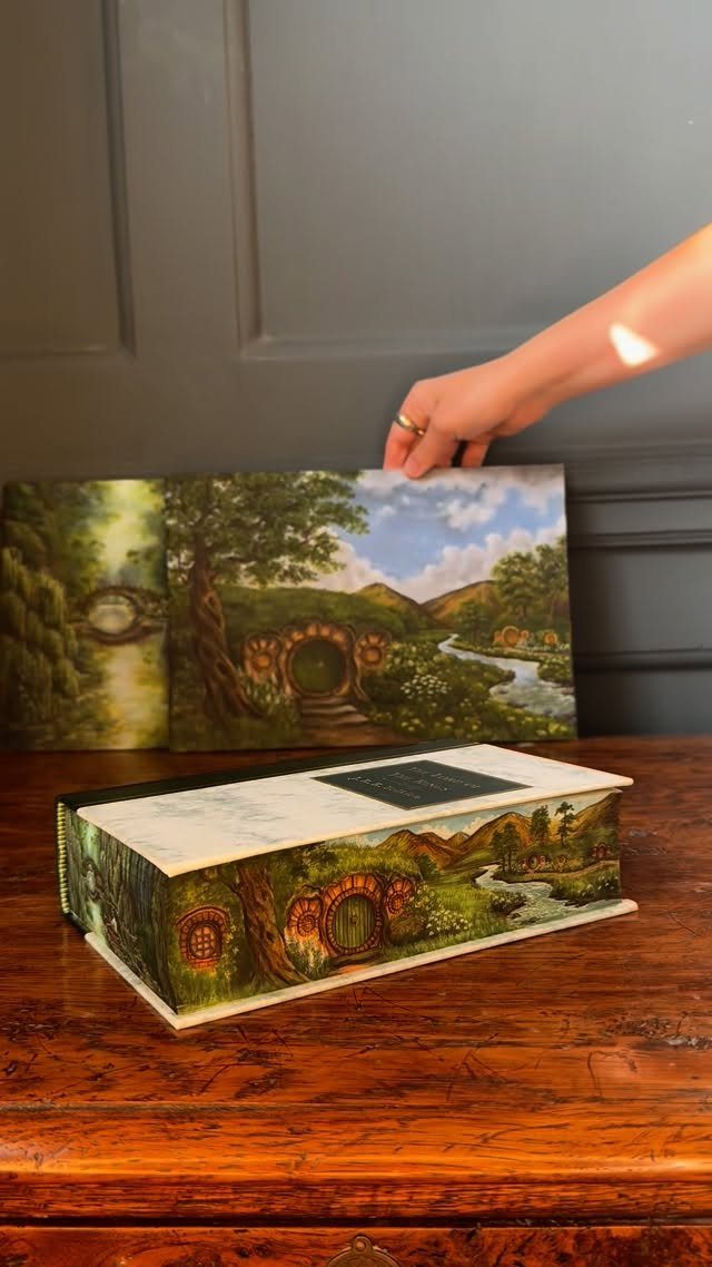 Hand holding an open book with a painted landscape of a hobbit-hole.