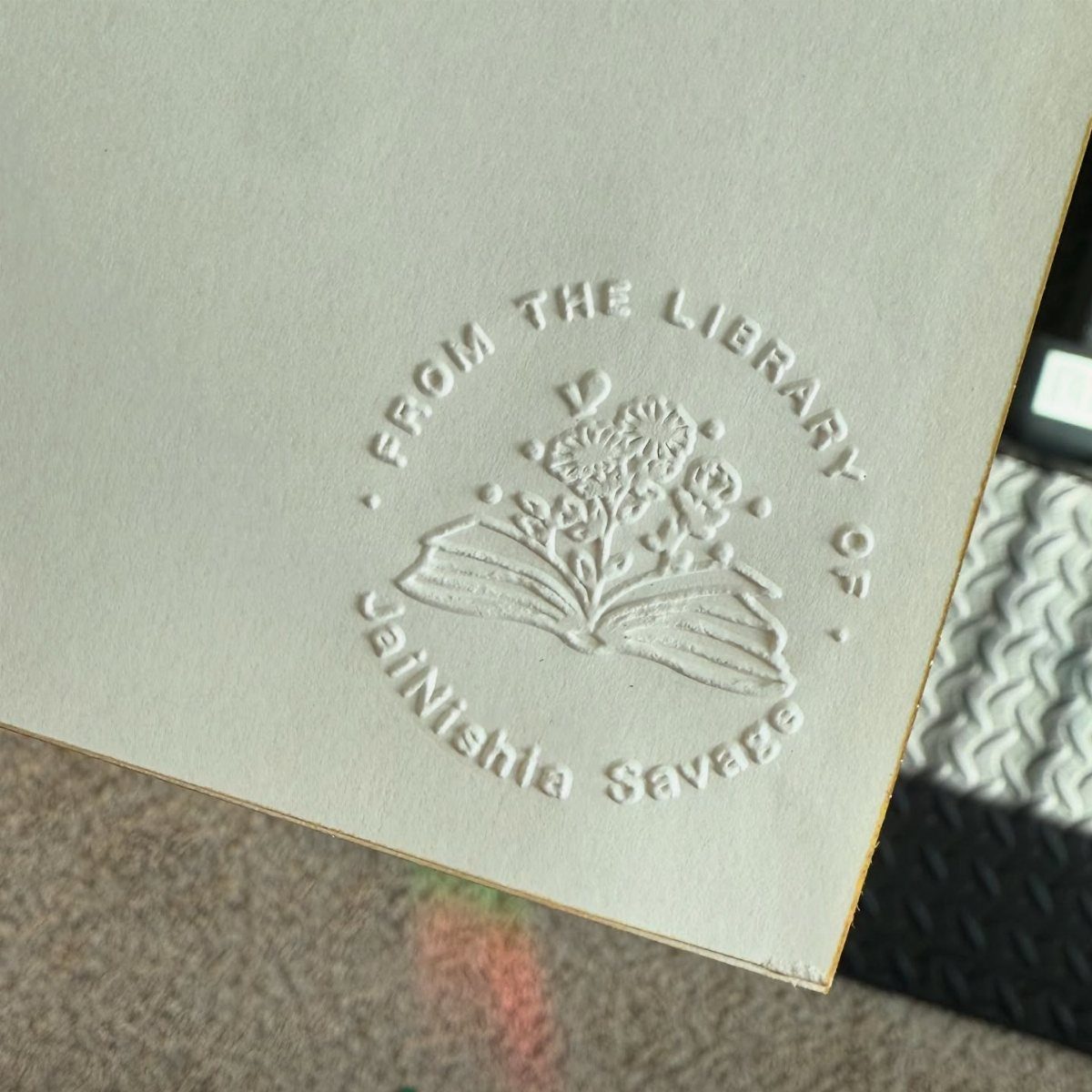 Embossed white bookplate featuring an open book with flowers and personalized name.