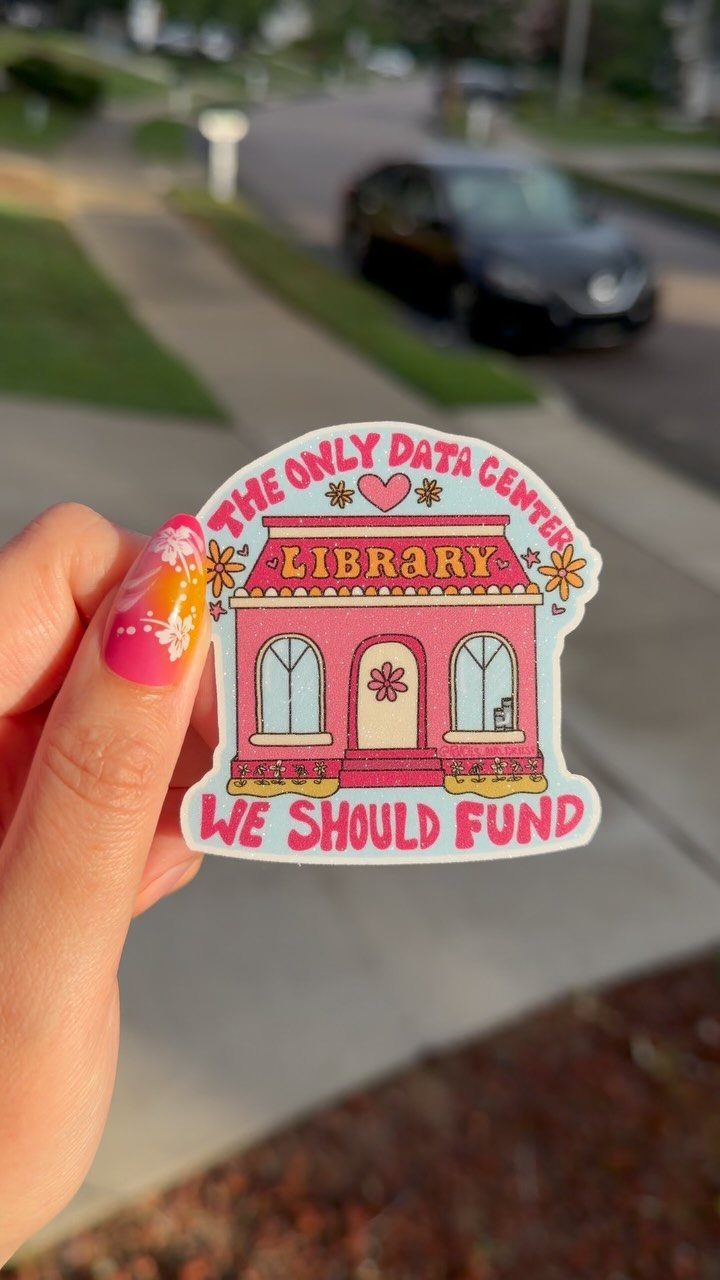 Pink library sticker held by a hand with colorful nails outdoors.