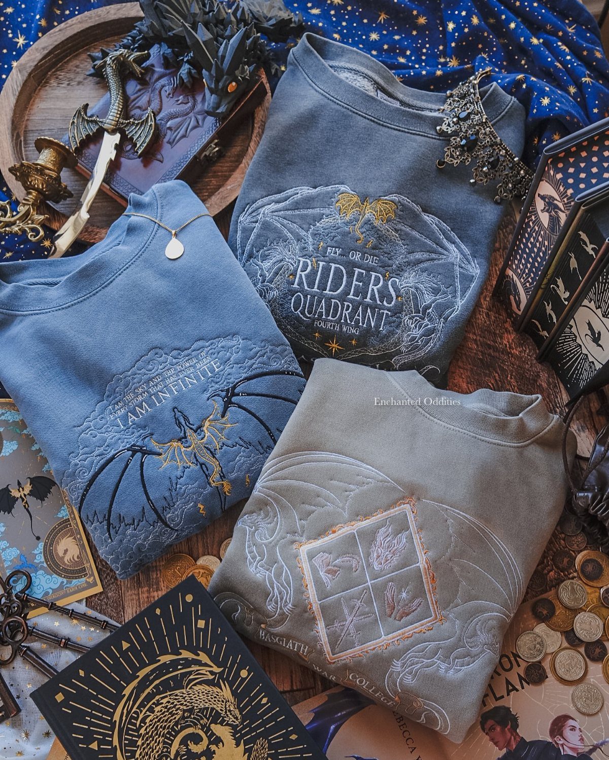 Three embroidered fantasy-themed sweatshirts surrounded by dragon figurines and gold keys on wood.