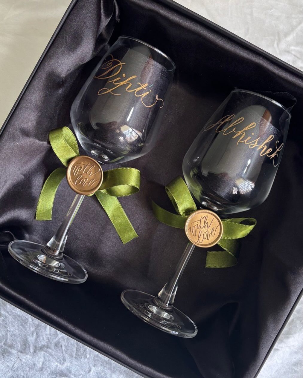 Two personalized wine glasses with gold names and green ribbons in a black box.