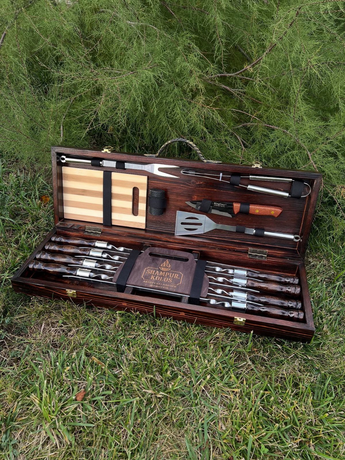 Barbecue tool set in a wooden case resting on green grass.