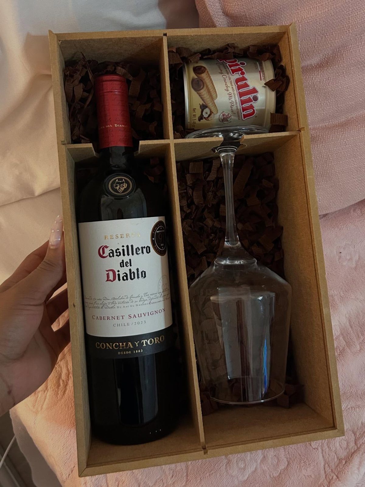 Gift box with red wine bottle, wine glass, and cookies in brown filler