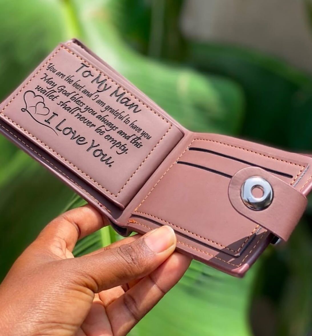 Brown leather wallet with an engraved sentimental message held against green foliage.