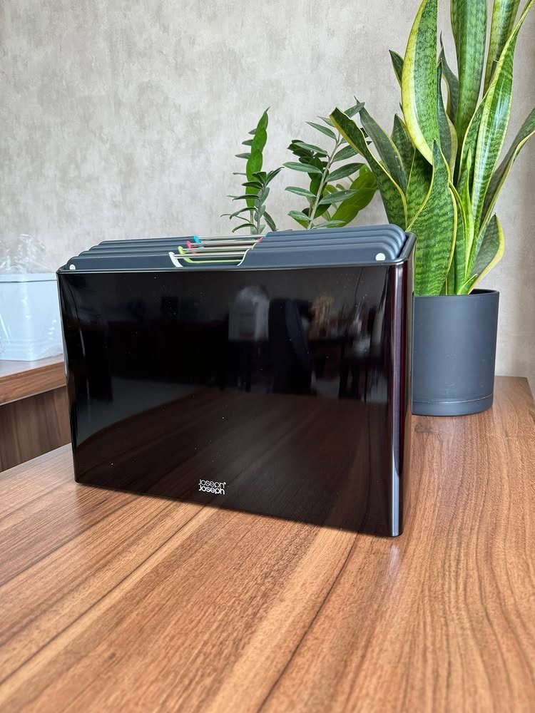 Black glossy bread box on a wooden table next to a snake plant.