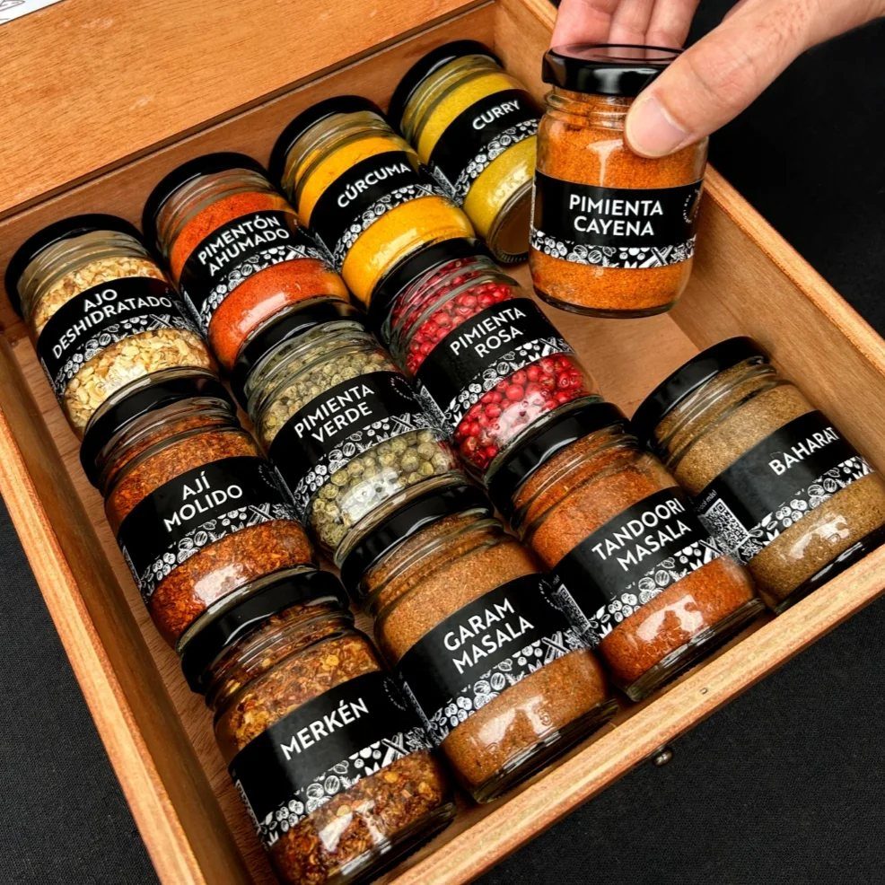 Assorted colorful ground spices in labeled glass jars inside a wooden storage box.