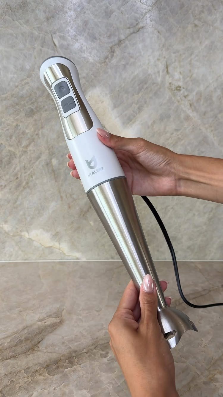 Immersion blender held by hands against a light marble countertop and wall background.