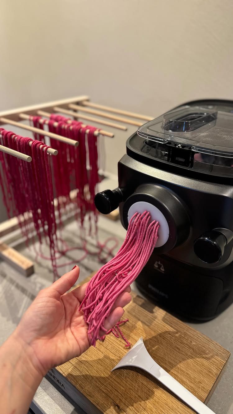 Black electric pasta maker extruding pink noodles into a hand with drying rack.