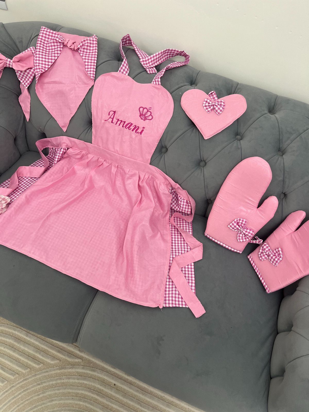 Pink personalized apron and matching kitchen accessories laid out on a grey sofa.