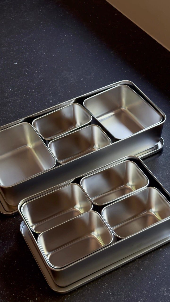 Stainless steel compartment organizers for spices or ingredients on a dark countertop.
