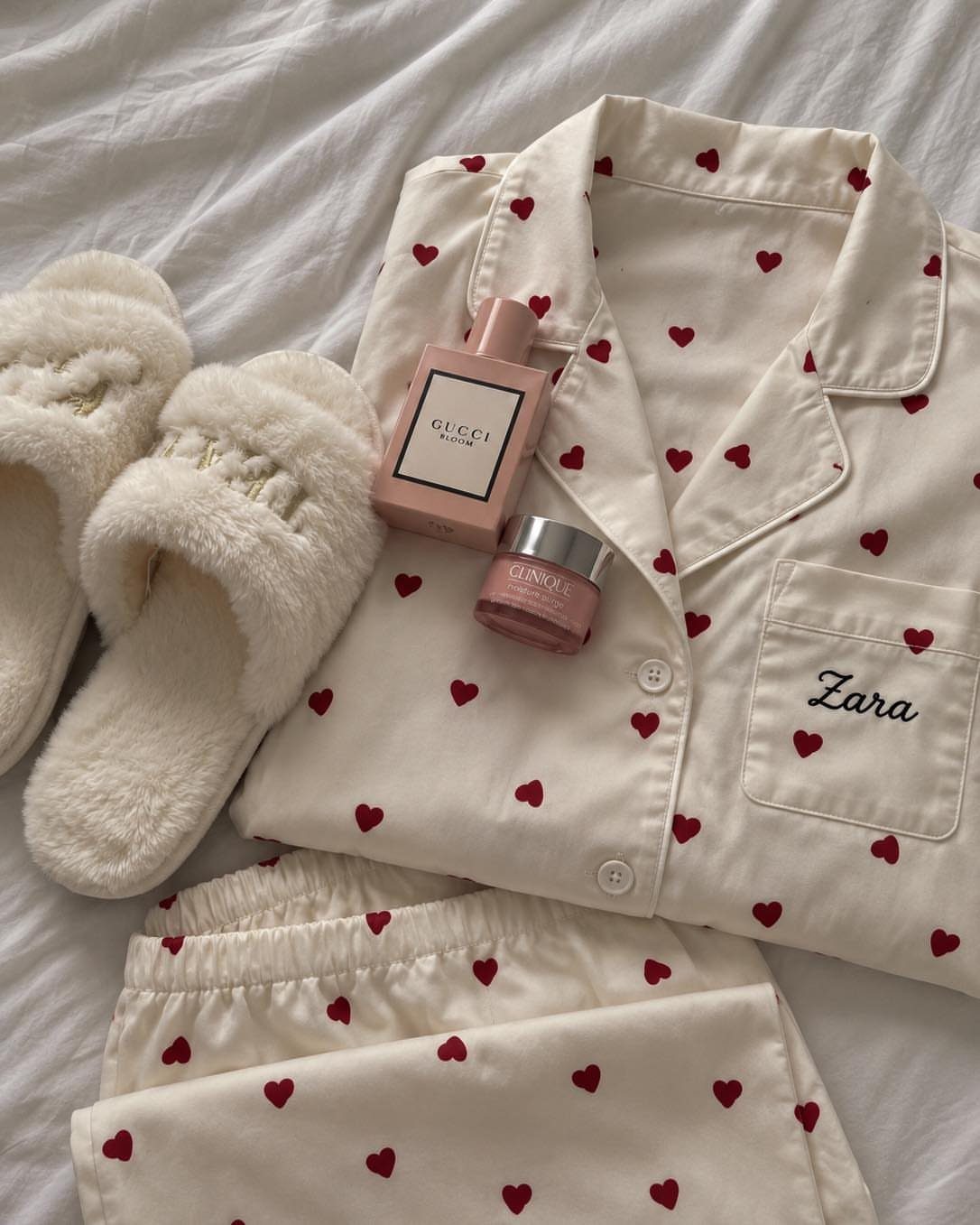 White heart-patterned pajama set with fluffy slippers and pink beauty products on white fabric.