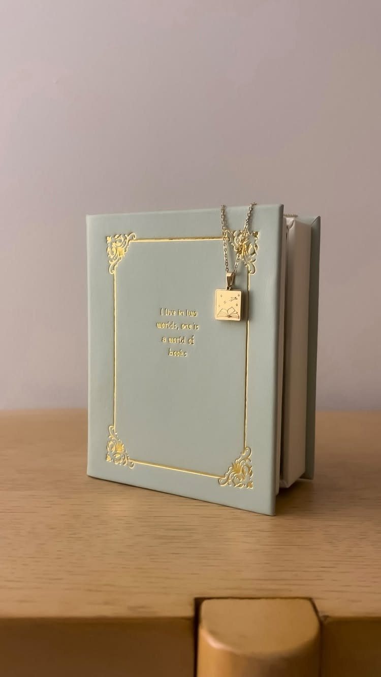 Gold square pendant necklace resting on a light blue book with gold detailing.