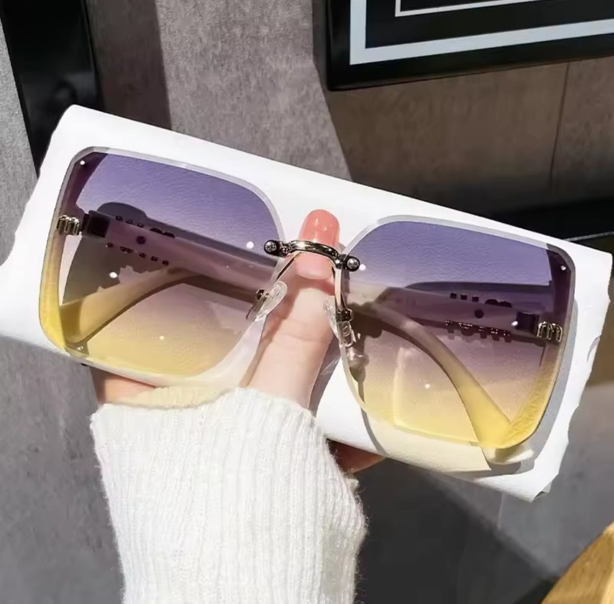 Oversized square sunglasses with purple and yellow gradient lenses held by a hand.