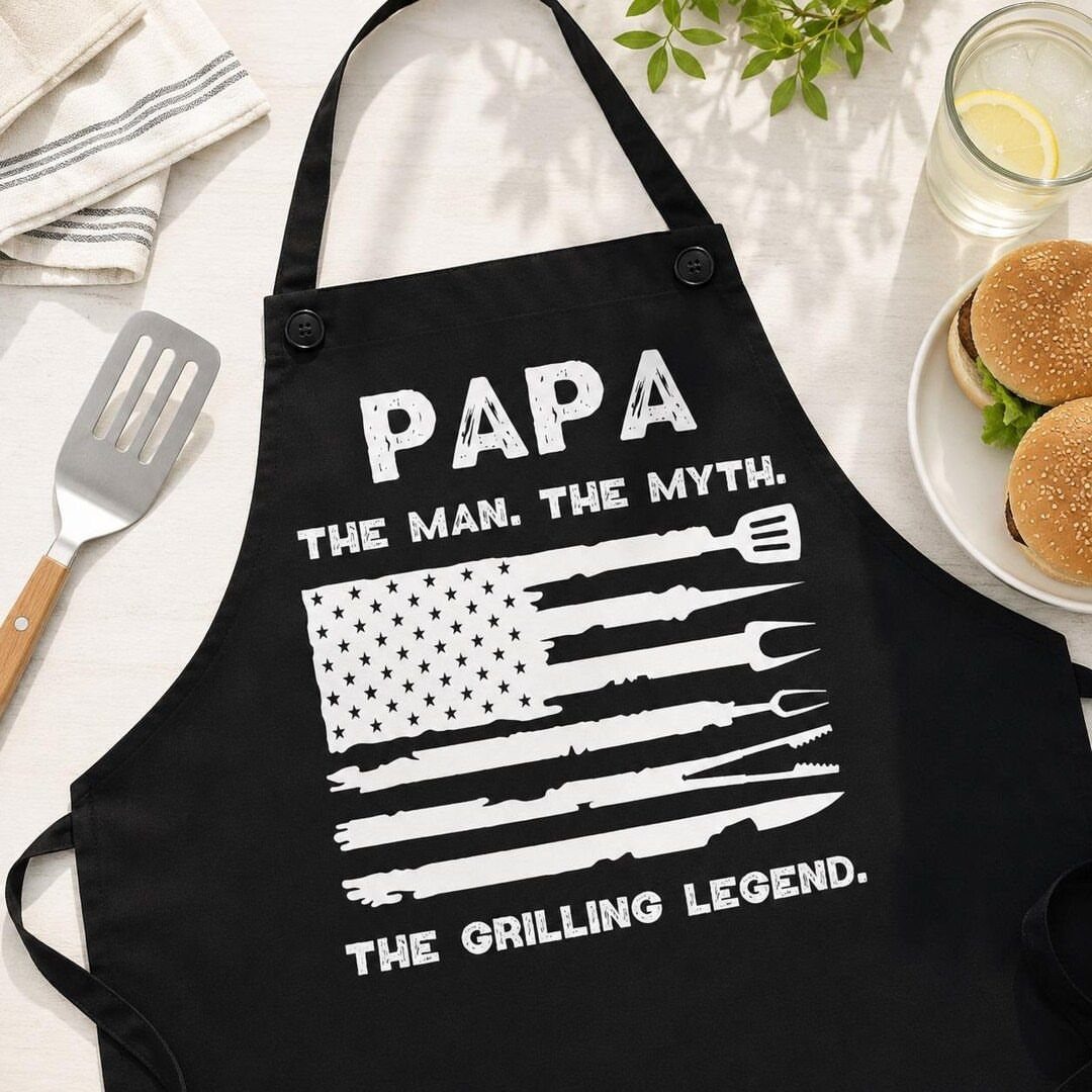 Black apron with Papa the Grilling Legend text and grilling tool flag graphic.