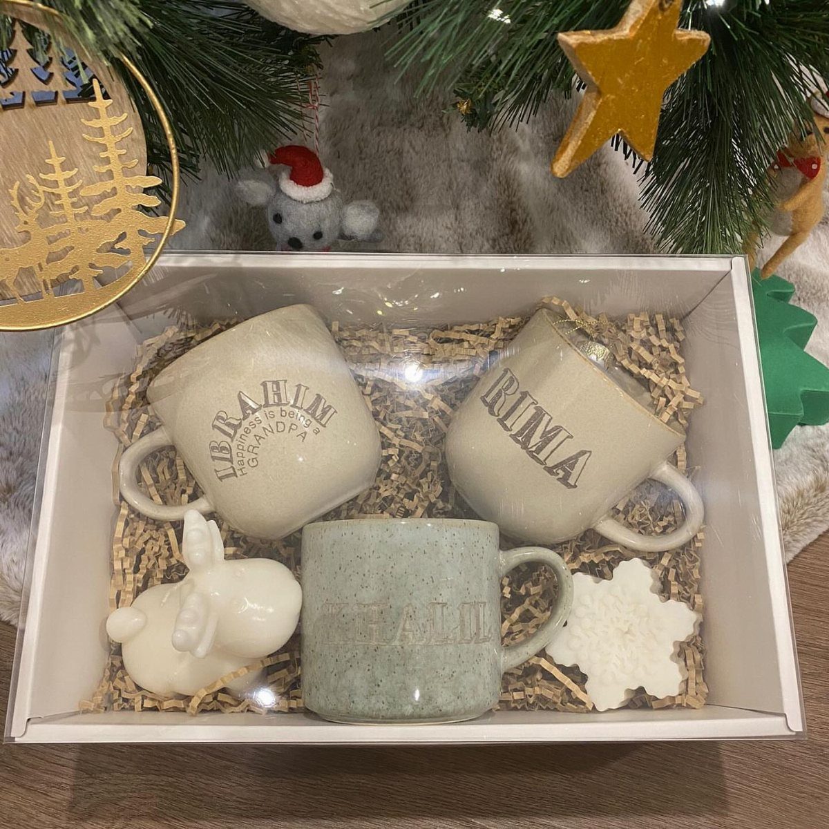 Three personalized ceramic mugs and ornaments in a white gift box with shredded paper.
