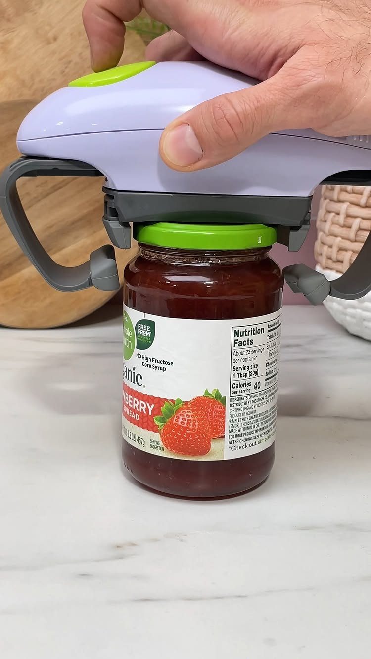 Electric jar opener removing the lid from a strawberry jam jar on marble.