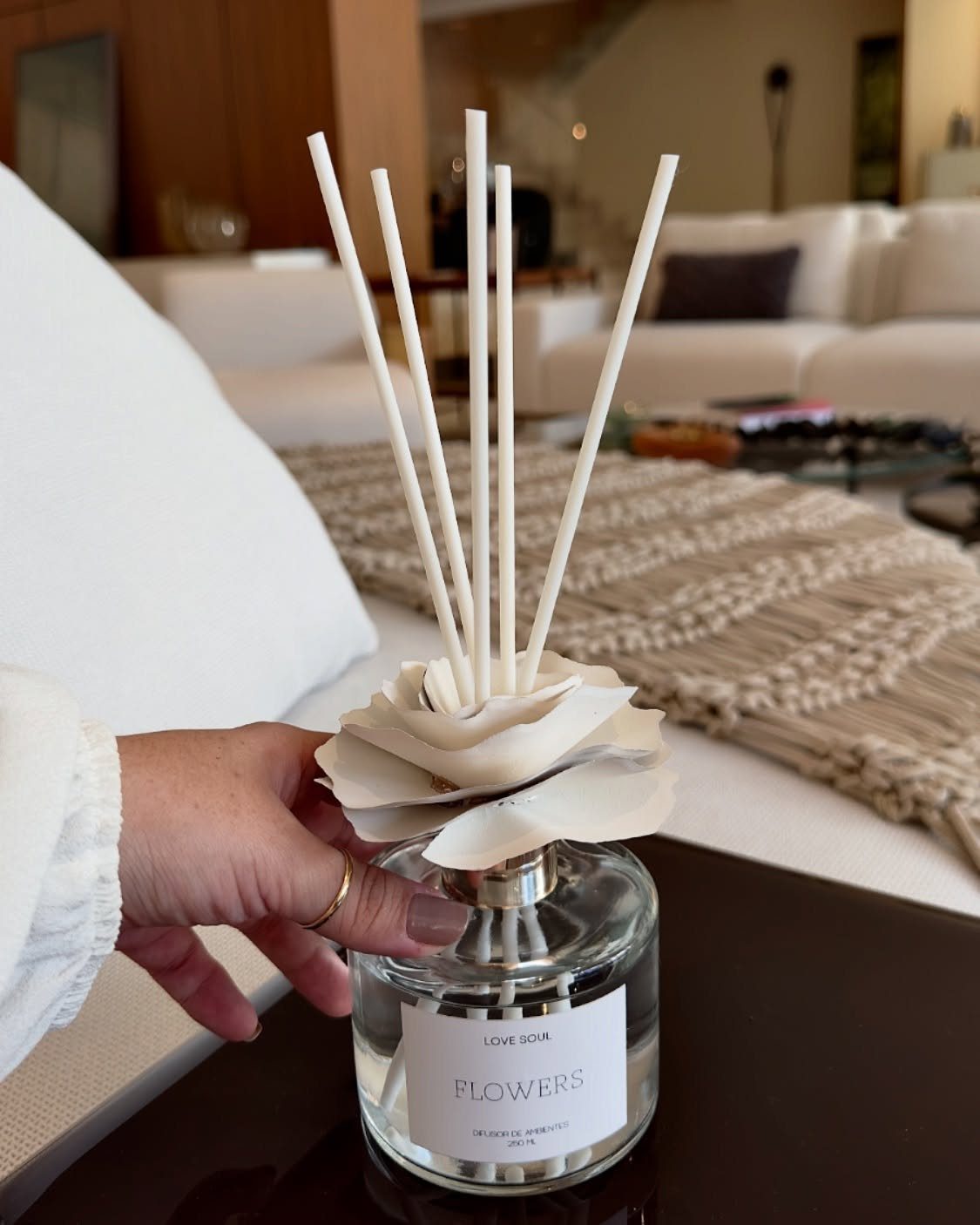 Fragrance diffuser bottle with white reeds and flower topper held by a hand.