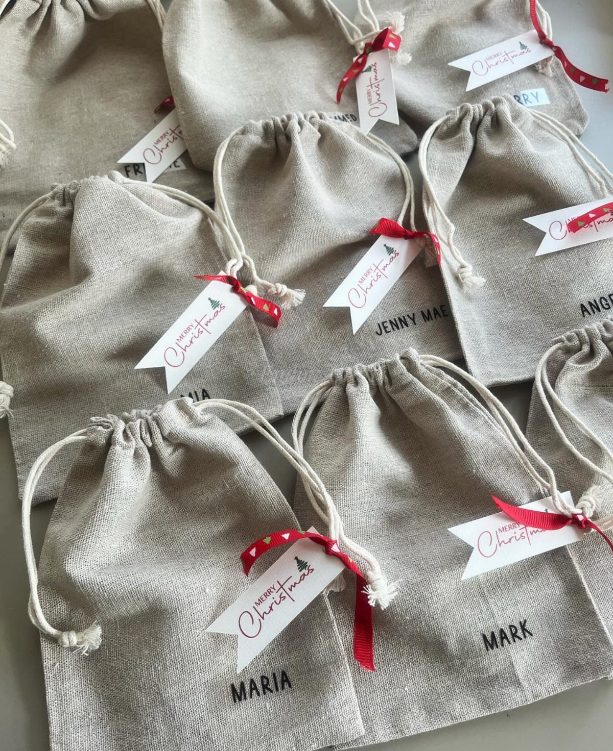 Beige linen drawstring gift bags with personalized names and Christmas tags.