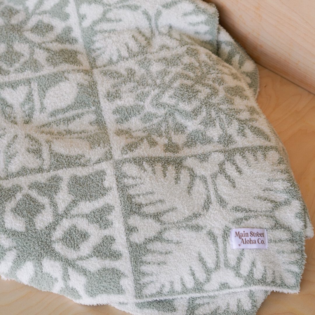 Sage green and white tropical leaf patterned plush blanket on a wooden surface