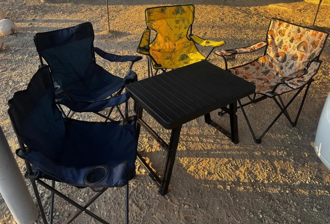 Folding camping chairs and a small black table set up on sandy ground.
