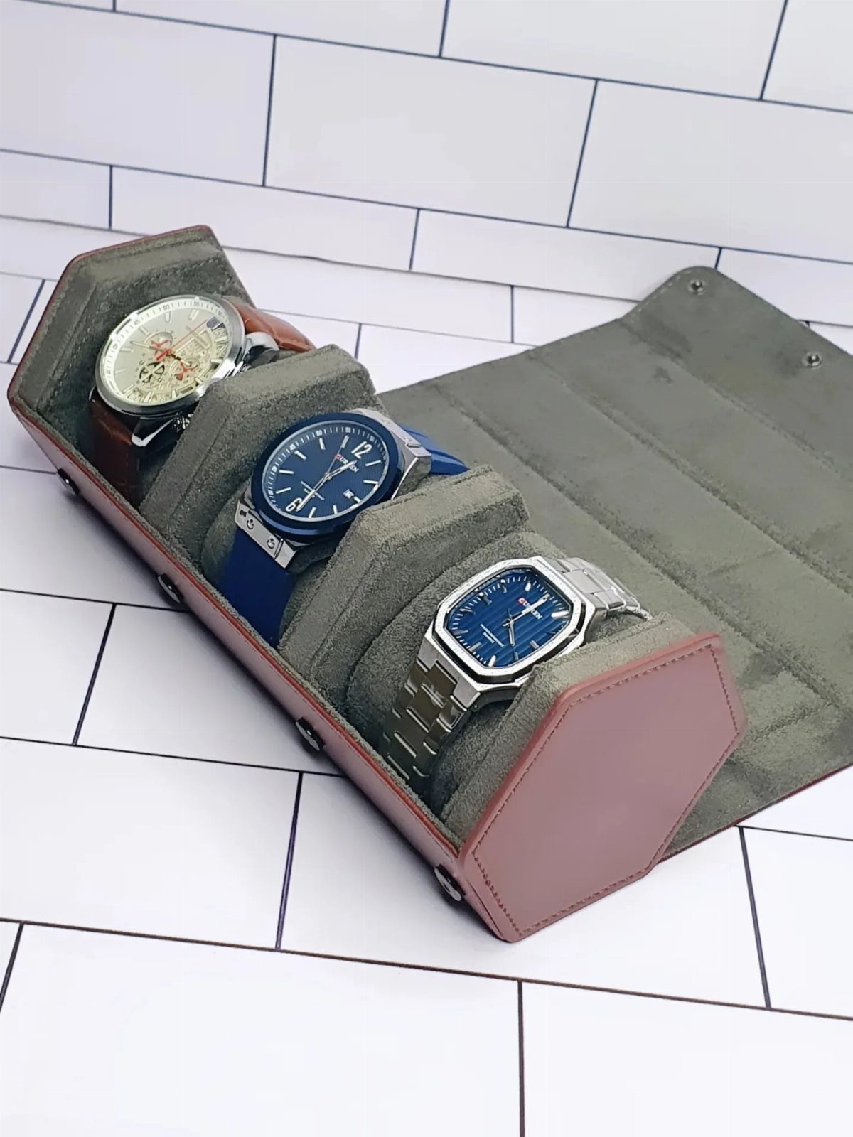 Three wristwatches stored inside a brown leather travel watch roll on white bricks.