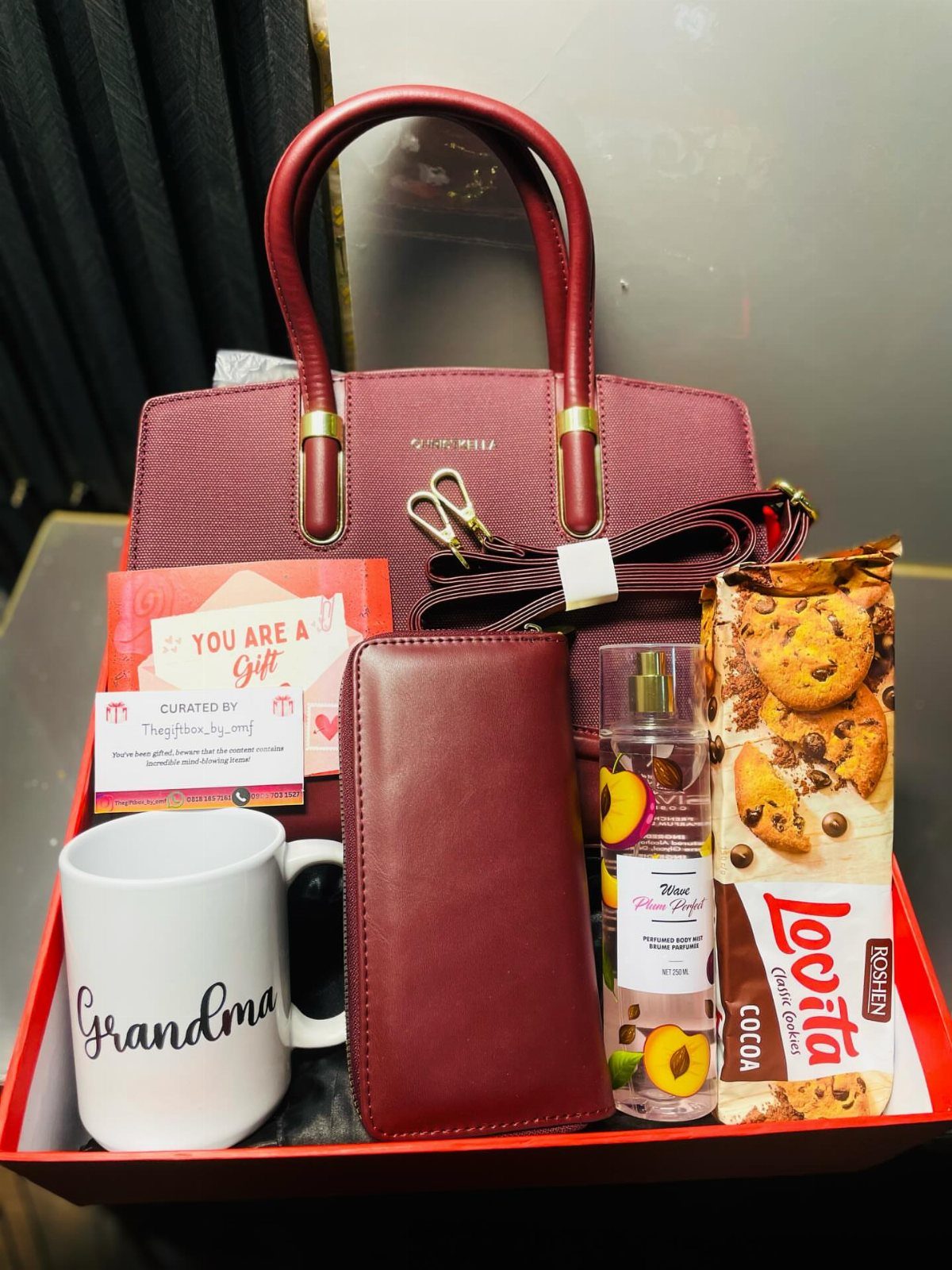 Red gift tray with handbag, wallet, Grandma mug, perfume, and chocolate cookies.