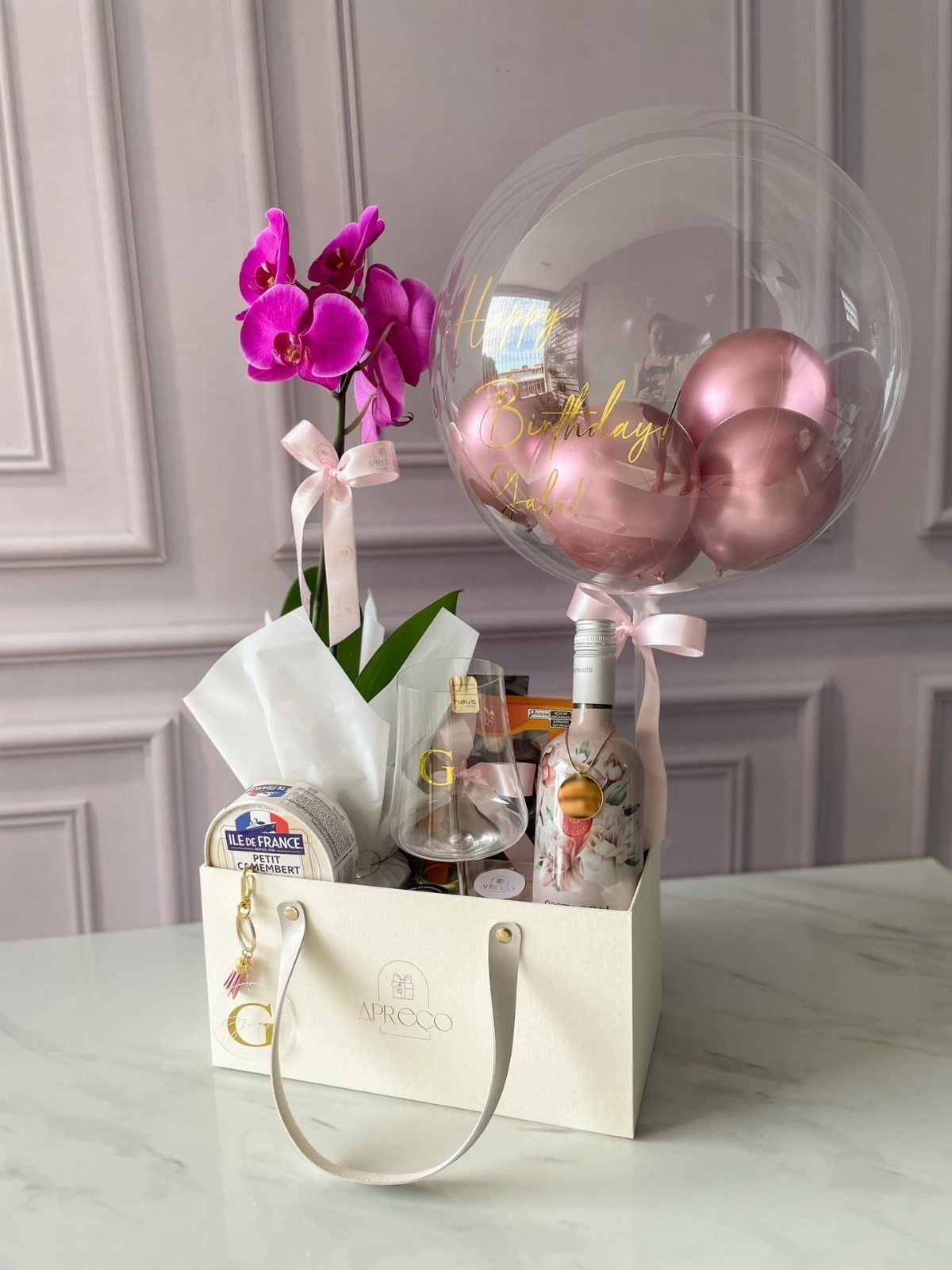 Gift bag with wine, a glass, purple orchid, and pink balloons on marble.