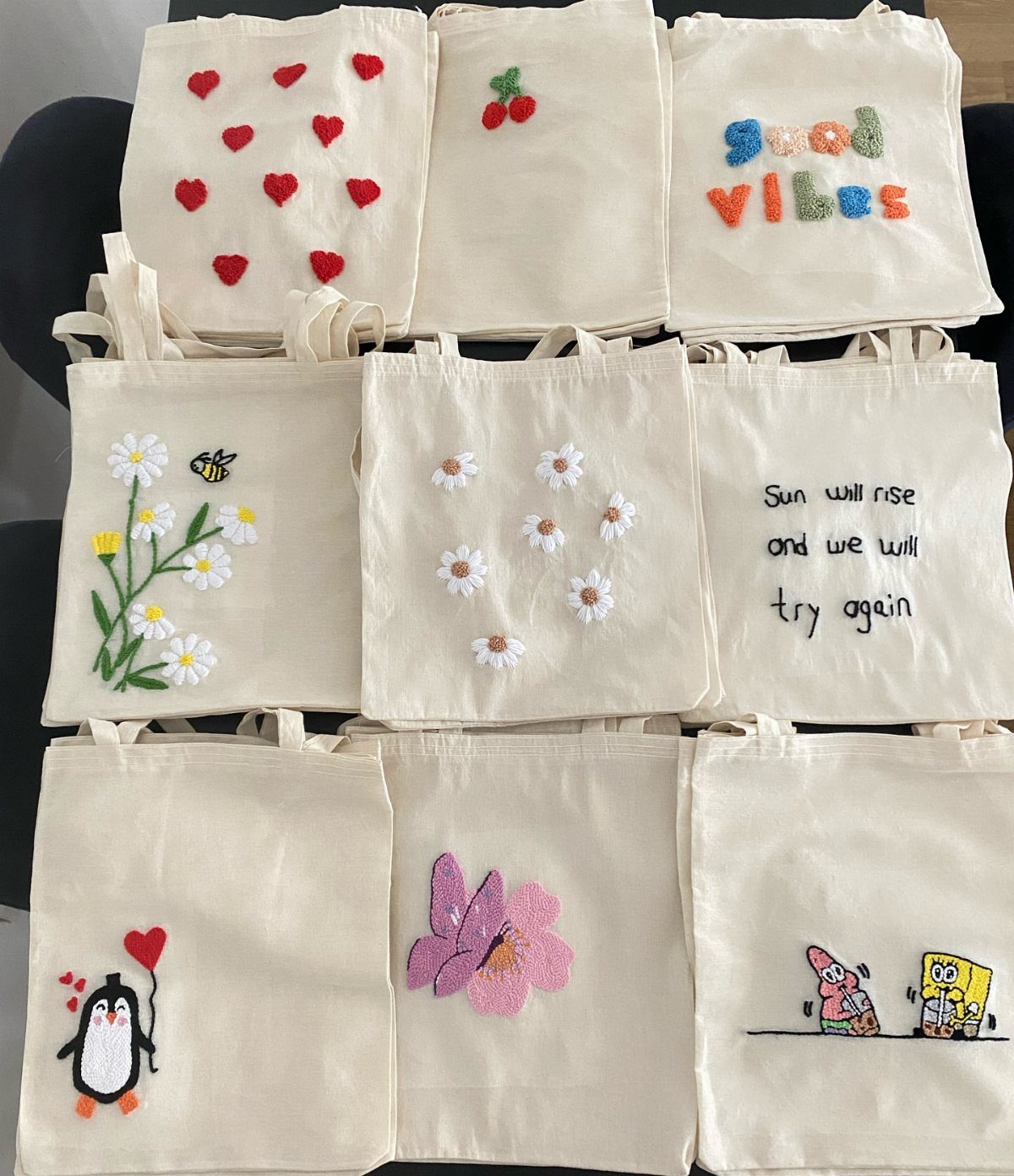 Nine cream canvas tote bags with various colorful embroidered designs and quotes.