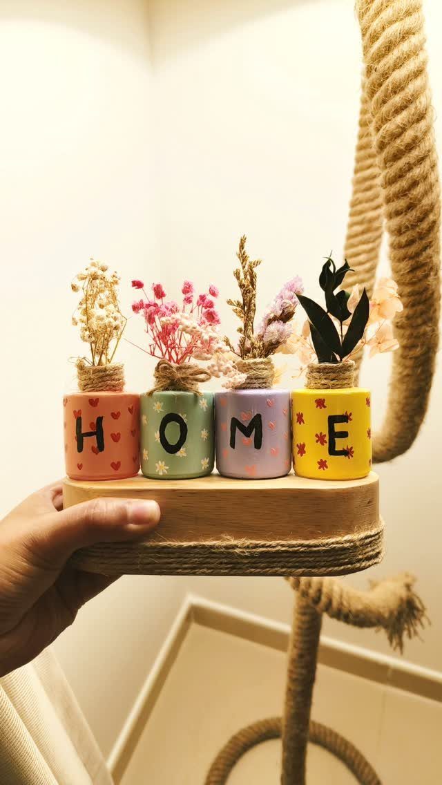 Four colorful miniature jars spelling HOME with dried flowers on a wooden tray.