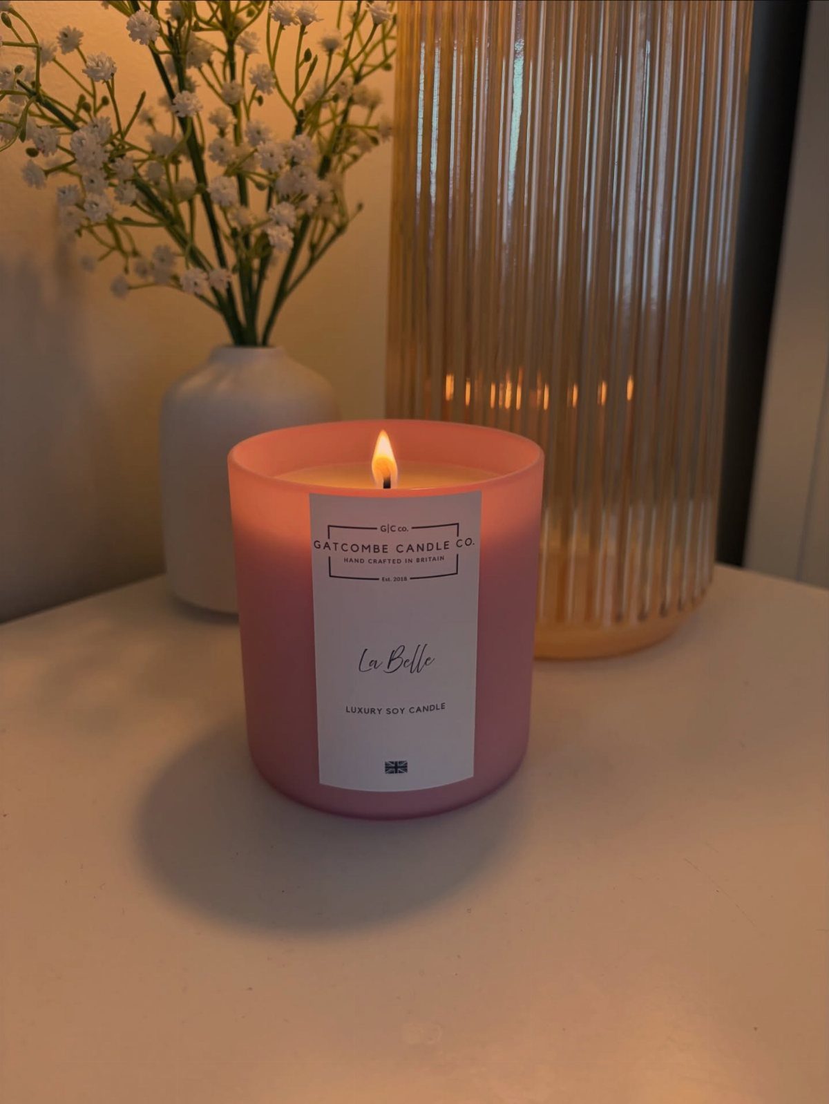 Lit pink scented candle on a white table next to white flowers and lamp