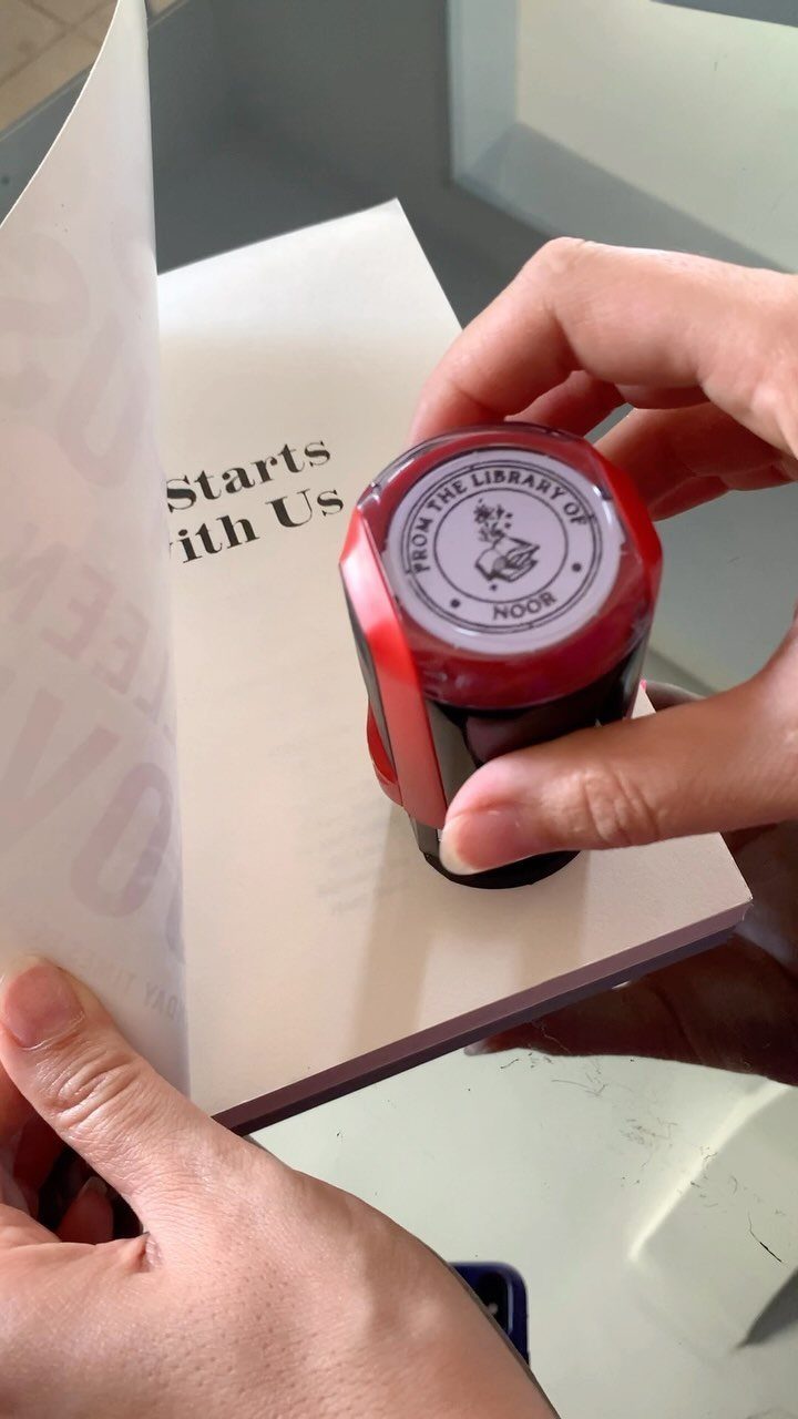 Hands holding a red personalized book stamp over an open white book page.