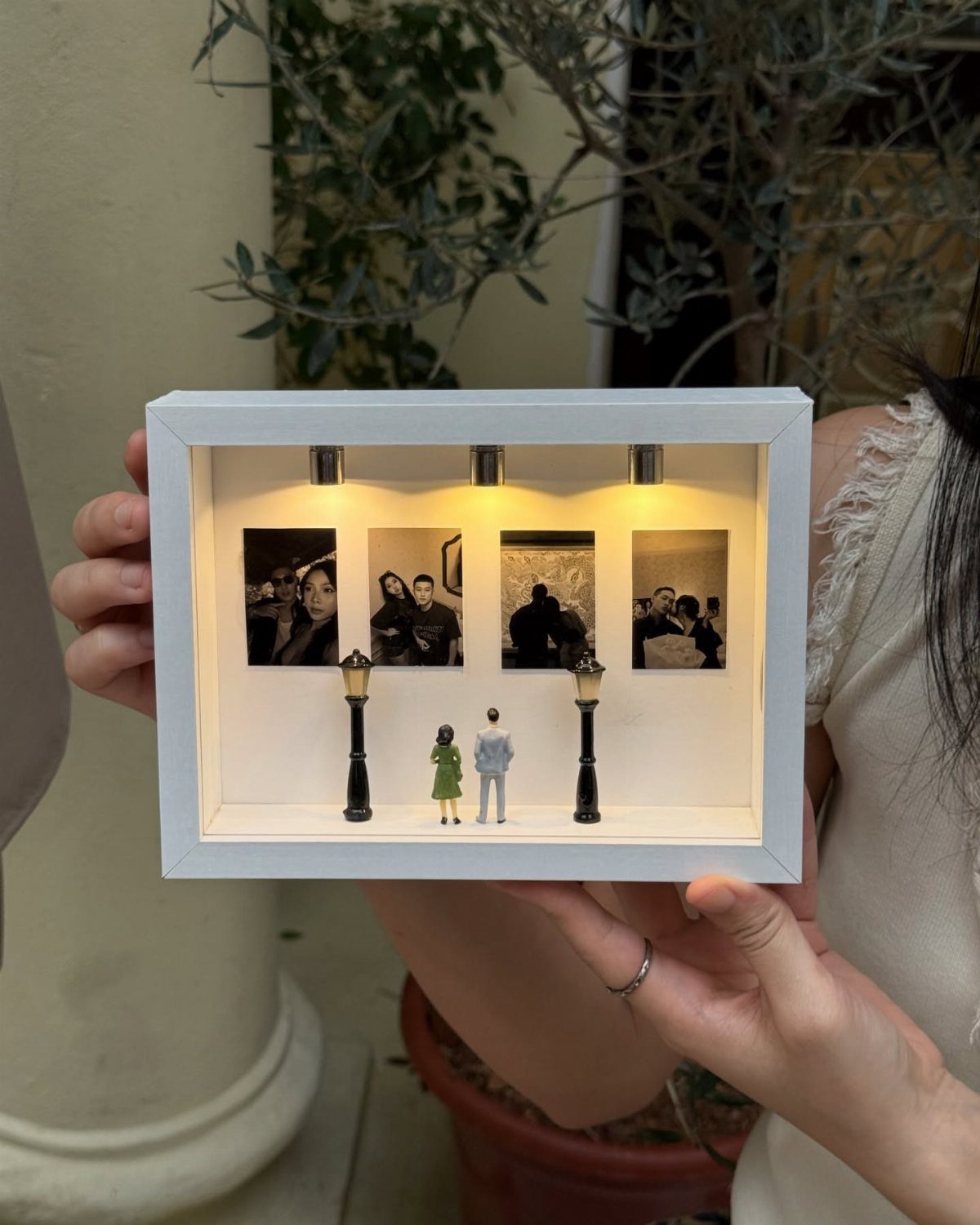 White shadow box gift with photos, miniature couple, and LED lights held by hands