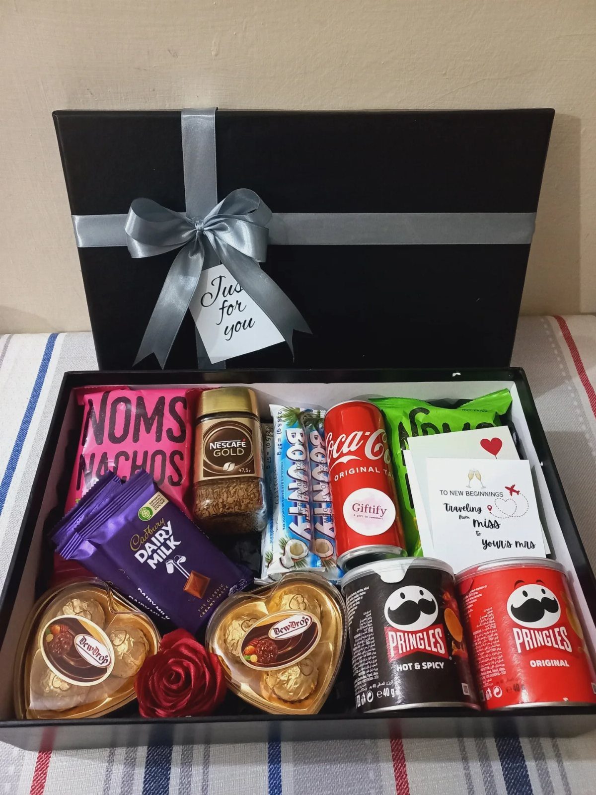 Black gift box filled with assorted snacks, chocolates, and drinks with silver ribbon.