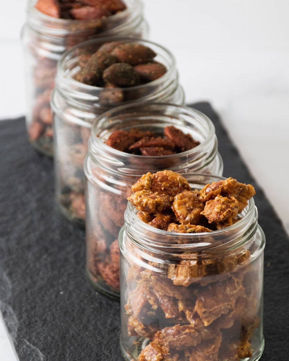 Four glass mason jars filled with spiced nuts on a dark slate surface.