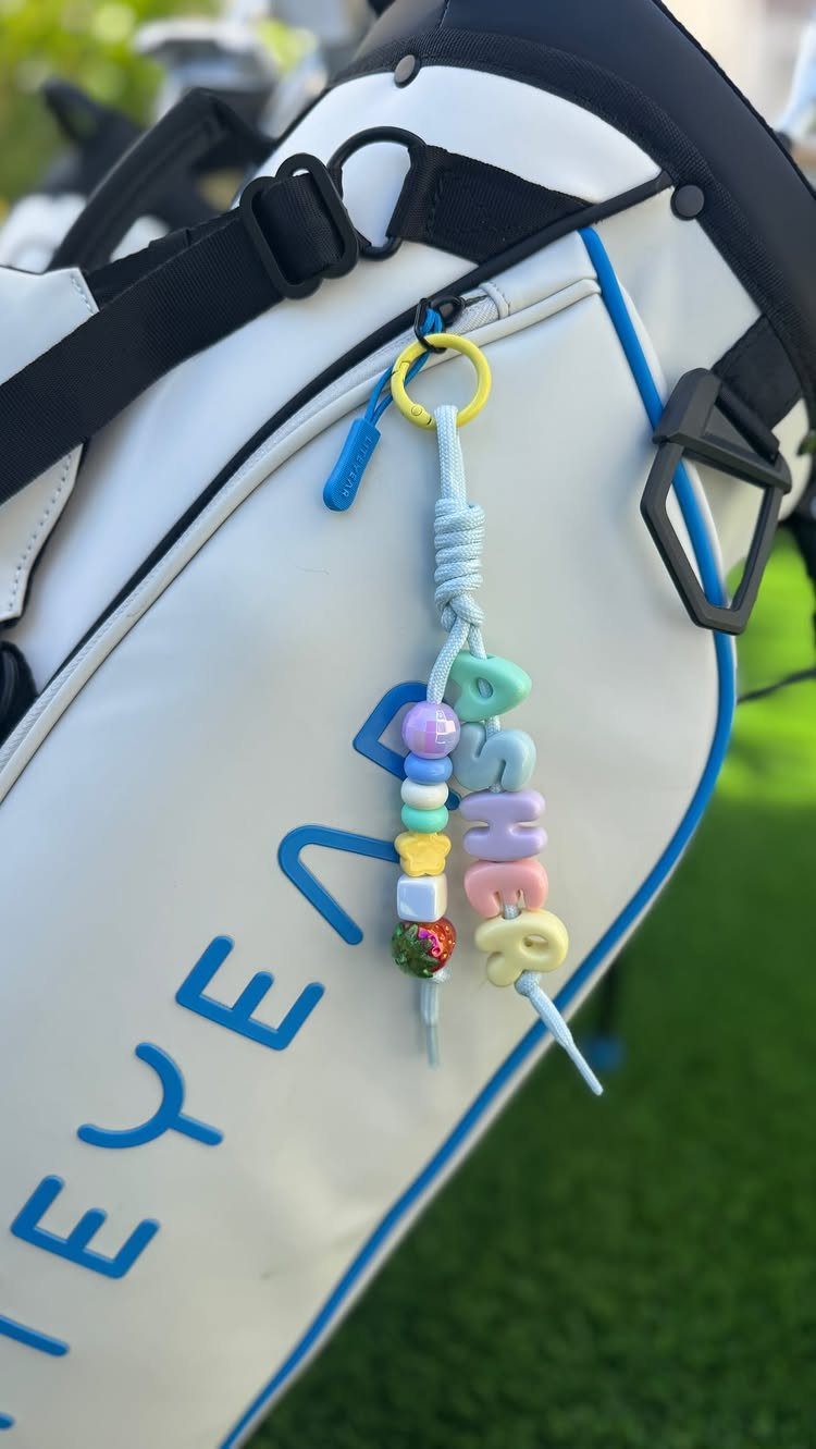 White golf bag with blue lettering and a colorful beaded name keychain attached.