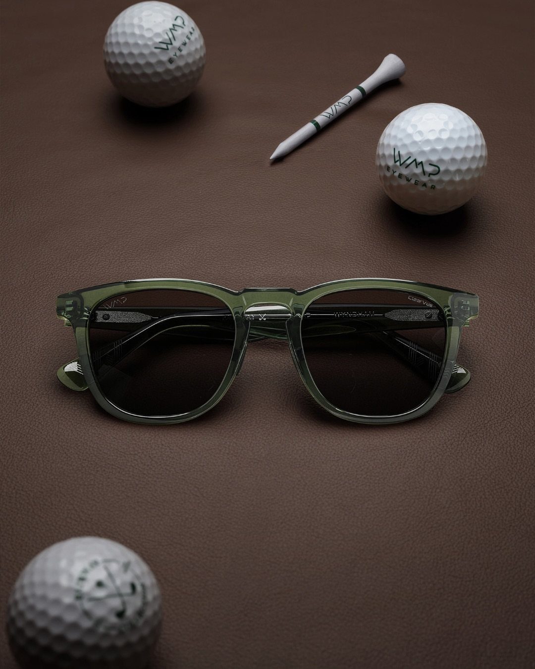 Transparent green sunglasses surrounded by white golf balls and a tee on brown leather.