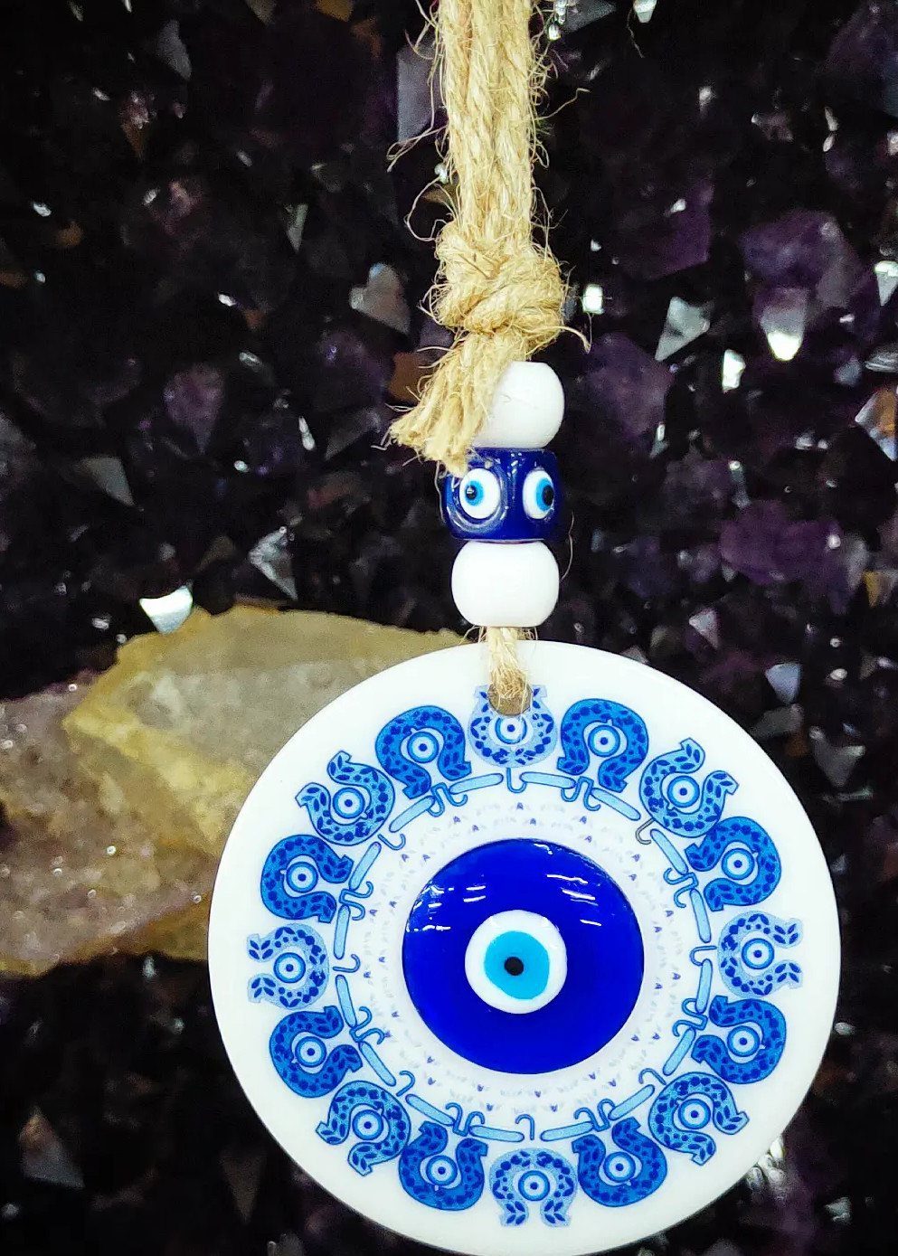 Evil eye ornament with blue patterns hanging from twine against purple crystals