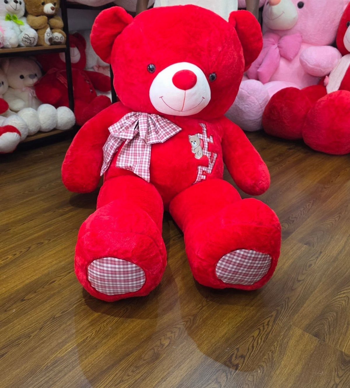 Large red plush teddy bear with a checkered bow on a wooden floor