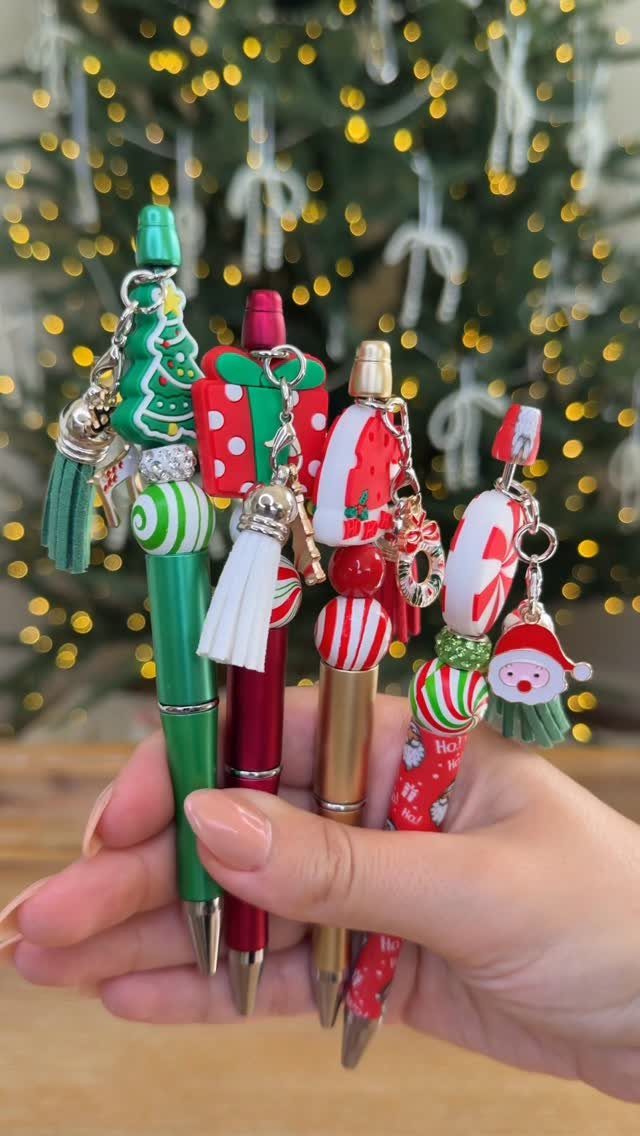 Four decorative holiday pens with Christmas charms held against a blurred lit tree.