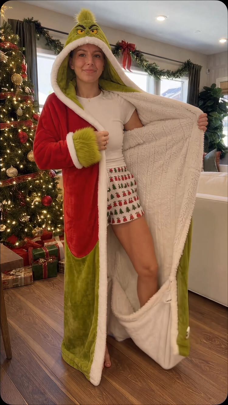 Woman wearing a Grinch-themed hooded robe standing beside a decorated Christmas tree.