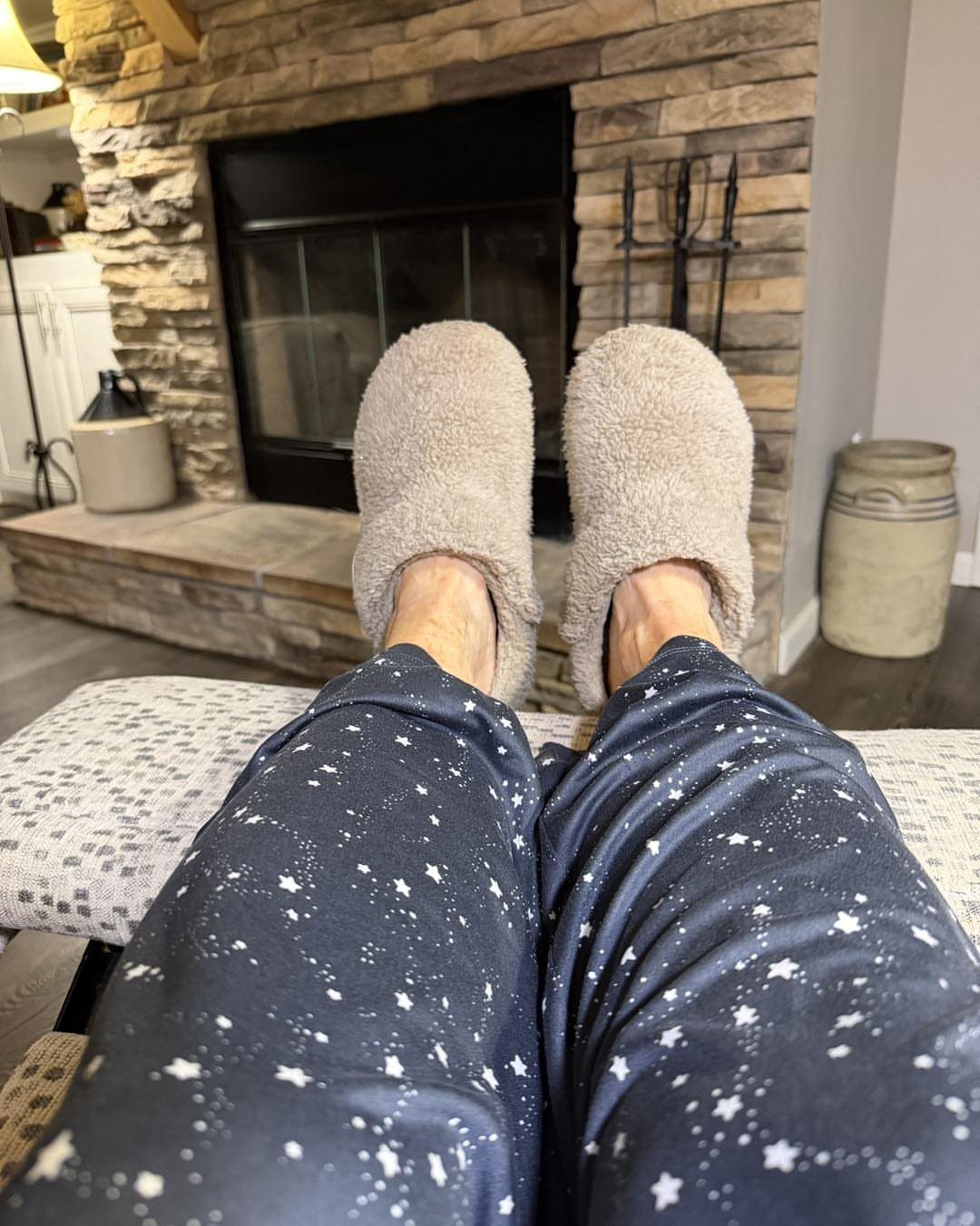 Beige fluffy slippers and star-patterned pajama pants resting in front of a stone fireplace.