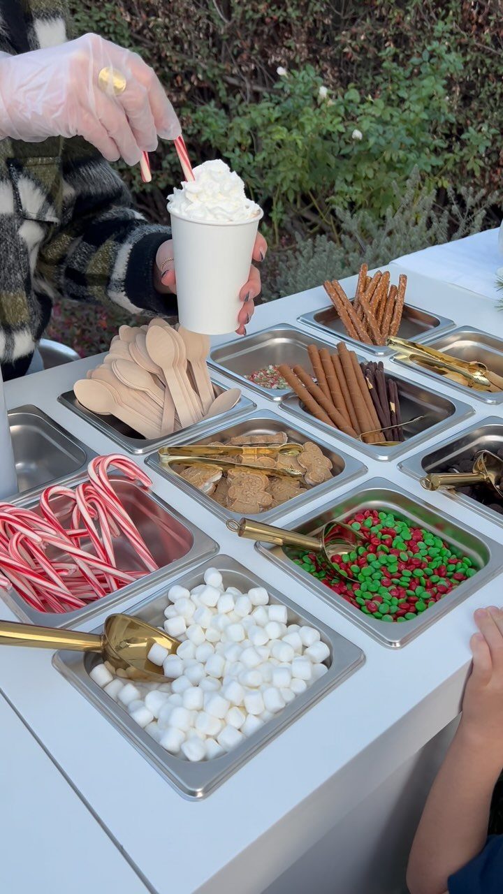 Hot cocoa with whipped cream and a variety of toppings in metal trays.
