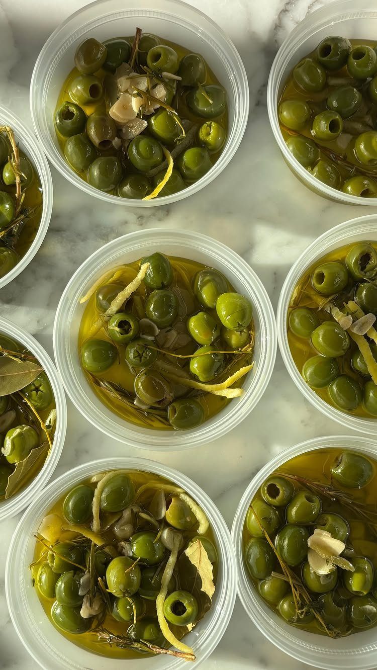 Marinated green olives with lemon zest in small clear plastic containers on marble.
