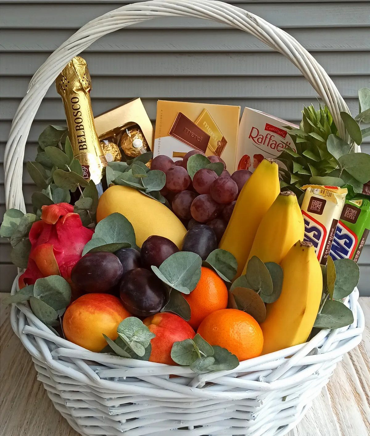 White wicker basket filled with fresh fruit, chocolate boxes, and sparkling wine.