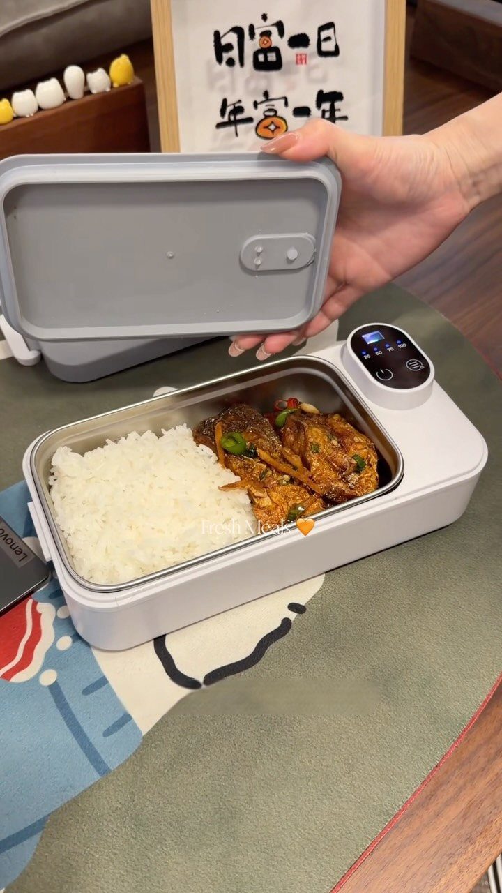 Electric heating lunch box with rice and meat being opened by a hand.