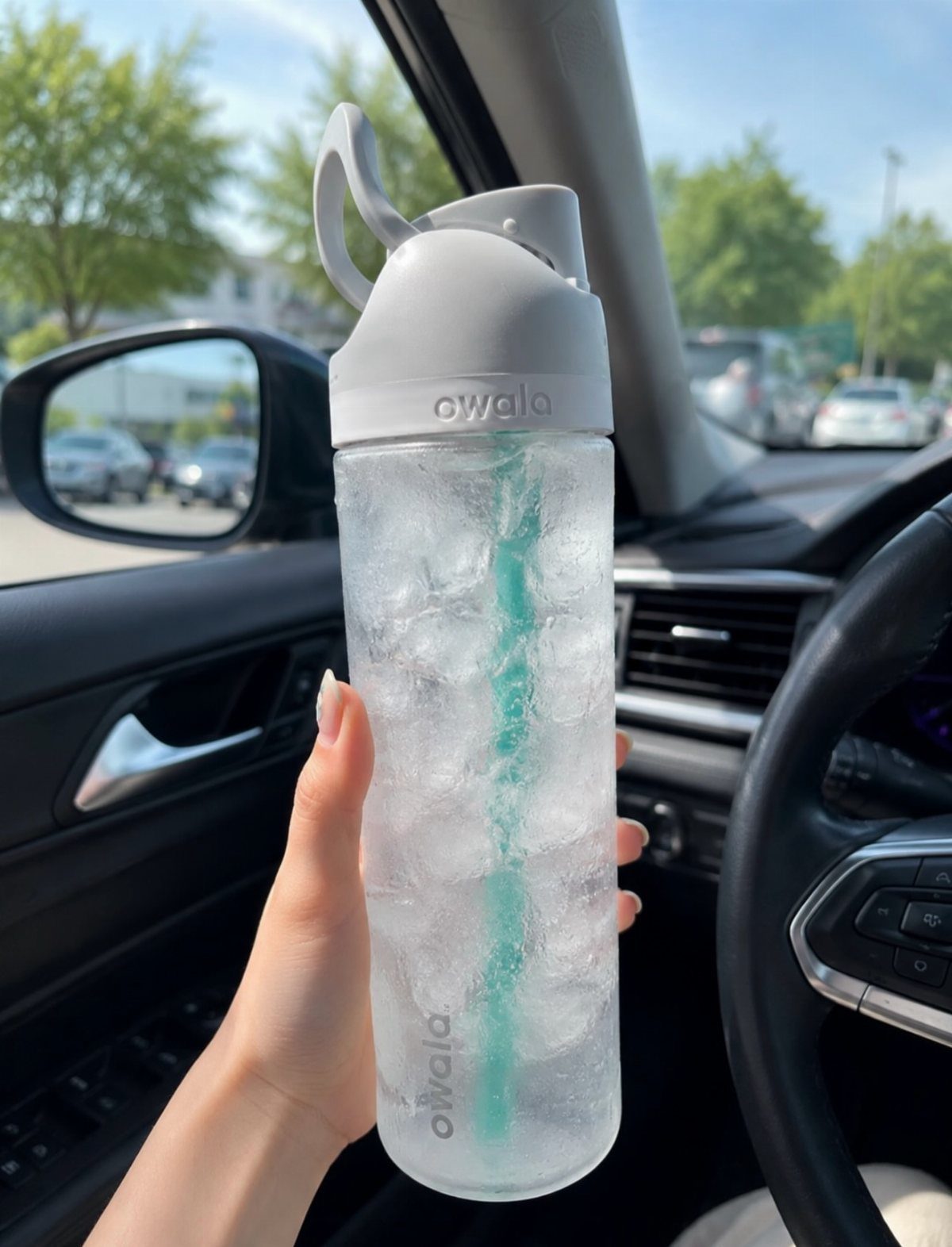 Clear Owala water bottle filled with ice and a teal straw held in a car